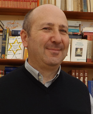 Ritratto in primo piano del Consigliere comunale Adriano Marini con maglione scuro e camicia chiara. Sullo sfondo una libreria.